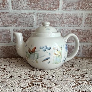 May include: White ceramic teapot with a lid and handle. The teapot is decorated with a rooster, blue flowers, and other whimsical illustrations. The teapot is sitting on a lace tablecloth.