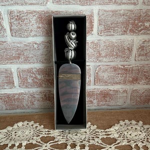 May include: A silver-toned cake server with a decorative handle featuring a cluster of stylized fruits and vegetables. The server is presented in a black box against a brick wall and lace doily.
