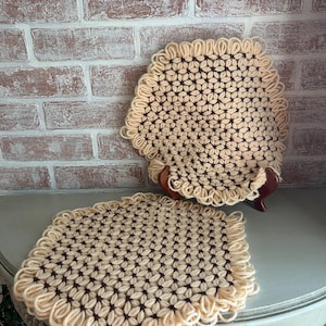 May include: Two handmade, beige and brown crochet trivets. One is rectangular, the other is round and displayed on a small wooden stand. The crochet pattern features a floral design with a fringed edge. The background is a brick wall.
