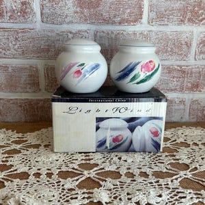 May include: Two white ceramic salt and pepper shakers with lids, decorated with floral and abstract designs in pink, blue, and green. They sit on a box that reads "Light Wind". The background is a brick wall.