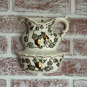 May include: Cream-colored ceramic wall-mounted pitcher and basin set. The set is decorated with green leaves, gold accents, and gold-colored grape clusters. The pitcher has a handle and a spout. The basin is below the pitcher.
