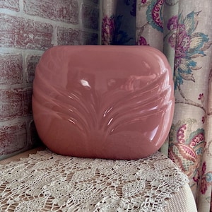May include: A glossy, dusty rose-colored ceramic vase with a rounded, flattened rectangular shape. The front surface features a symmetrical, molded fan-like pattern of raised, curved ridges that radiate outward from the center base.