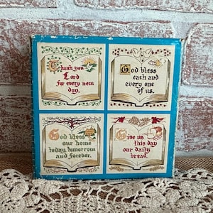 May include: A square, blue-bordered decorative item with four panels, each featuring religious text and illustrations. The panels include phrases like "Thank you Lord" and "God bless us," with images of open books and decorative elements.