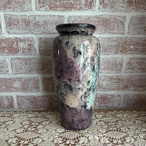 May include: A tall, decorative vase with a speckled, glazed finish. The vase features a dark rim and a body with shades of purple, black, and light blue. The vase is set against a brick wall and a lace tablecloth.