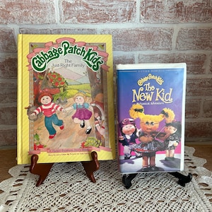 May include: Two Cabbage Patch Kids items: a book titled "The Just-Right Family" and a VHS tape titled "The New Kid Musical Adventure". Both items feature illustrations of Cabbage Patch Kids characters.