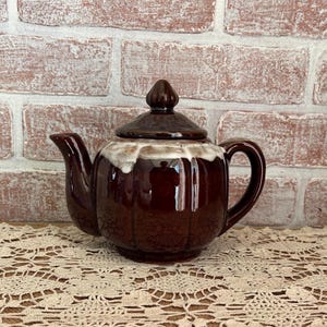May include: A brown ceramic teapot with a lid, spout, and handle. The teapot has a glossy finish and a white and brown rim. The teapot is sitting on a cream-colored lace tablecloth in front of a brick wall.