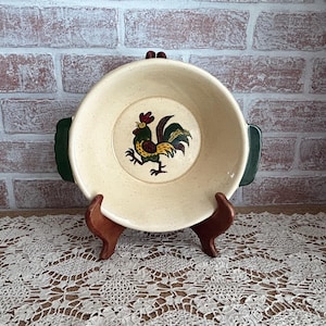 May include: Cream-colored ceramic dish with a rooster illustration in the center. The dish has green handles and is displayed on a wooden stand. The rooster is depicted in shades of red, green, yellow, and black.