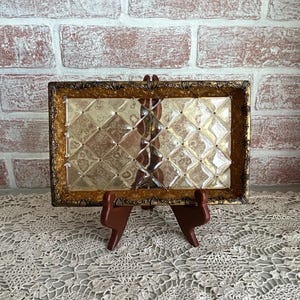 May include: A rectangular decorative tray with a gold-colored ornate frame. The tray's interior features a clear, textured glass surface with a diamond pattern. The tray is displayed on a brown wooden stand, set against a brick wall.