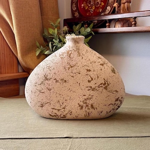 May include: A wide, rounded ceramic vase with a textured, off-white surface featuring irregular, mottled brown and gold-toned speckles. The vase has a narrow, circular opening at the top and a flattened, oval-shaped body that tapers toward the neck.