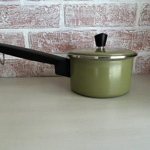 May include: A small, olive green saucepan with a black handle and lid. The handle has a metal loop for hanging. The lid has a black knob. The pot is set against a brick wall.