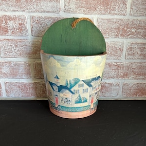 May include: A decorative wall pocket with a green wooden back and a cylindrical container. The container features a painted scene of a town with buildings, a blue sky, and a pink rim. A rope loop is attached for hanging.