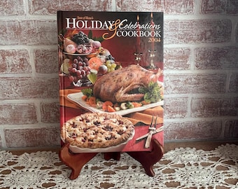 May include: A cookbook titled "Holiday Celebrations Cookbook 2004" is displayed on a wooden stand. The cover features images of a roasted turkey, fruit, and a pie. The background is a red and white brick wall.