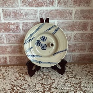 May include: A cream-colored ceramic plate with blue bamboo and floral designs. The plate has a central hole and is displayed on a dark brown wooden stand. The background features a brick wall.