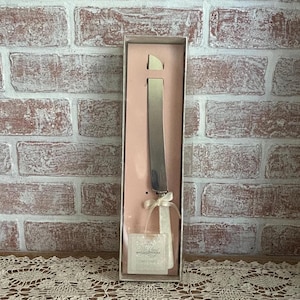 May include: A silver cake knife with a ribbon and tag, presented in a pink and beige box. The knife is displayed against a backdrop of a brick wall.
