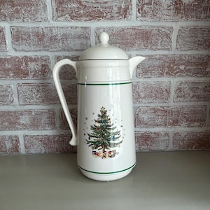 May include: White ceramic Christmas-themed coffee pot with a lid and handle. The pot features a green band and a Christmas tree design with presents. The pot is approximately 10 inches tall.