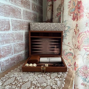 May include: A brown leather-textured backgammon set in an open case with a gold-tone metal latch. The interior features a dark brown and tan point design, white and dark brown checkers, dice, and a white cylindrical container labeled The Traveler.