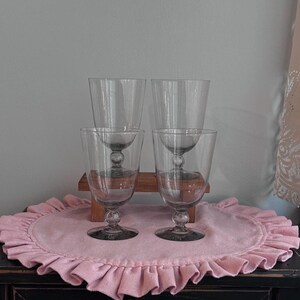 May include: A set of four clear glass goblets with a smooth, tapered bowl and a distinct spherical node on the stem. Each glass features a circular base and a simple, transparent design, arranged in two rows on a small wooden stand.