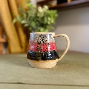 May include: A ceramic mug with a textured, light beige base and a handle. The exterior features a horizontal color gradient with a glossy black bottom section, a vibrant red middle band, and a light grey top, accented by a black tree illustration.