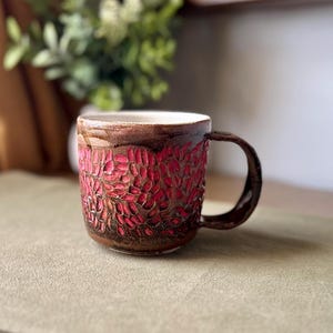 May include: A ceramic mug with a dark brown glazed exterior featuring a textured pattern of carved, vibrant pink leaf-like shapes. The mug has a smooth white interior and a curved handle on the right side, showcasing a handcrafted, rustic aesthetic.