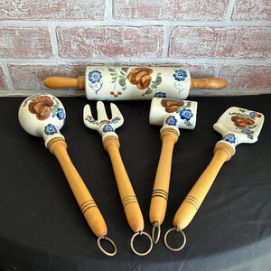 May include: A collection of ceramic kitchen tools with wooden handles. The set includes a rolling pin, a spatula, a fork, and two other tools. Each piece is decorated with a floral pattern in shades of brown, blue, and green.