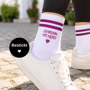 Könnte beinhalten: Weiße Socken mit zwei dunkelvioletten Streifen am oberen Rand. Der Text LEHRERIN MIT HERZ ist in Violett auf der Seite eingestickt, ergänzt durch ein kleines violettes Herzsymbol. Die Socken werden zu weißen Sneakern und schwarzen Leggings getragen.
