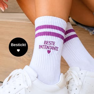 Könnte beinhalten: Weiße Socken mit zwei violetten Streifen am oberen Rand und dem gestickten violetten Schriftzug BESTE PATENTANTE mit einem kleinen Herzsymbol unter dem Text. Die Socken haben eine gerippte Struktur und werden mit weißen Sneakern getragen.