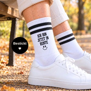 Könnte beinhalten: Weiße Crew-Socken mit zwei schwarzen Streifen am oberen Rand und dem schwarz gestickten Text "ICH BIN JETZT IN RENTE" über einem kleinen schwarzen Smiley-Symbol. Die Socken werden zu weißen Sneakern und einer hellen Hose mit Umschlag getragen.