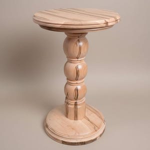 May include: A light-colored wooden side table with a round top and base, connected by a turned, baluster-style pedestal. The wood grain is visible, with darker streaks adding detail. The table is approximately 30 inches tall.