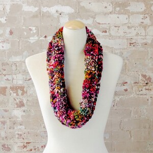 May include: A colorful knitted infinity scarf with a variety of shades of pink, red, orange, yellow, green, and blue. The scarf is displayed on a white mannequin.