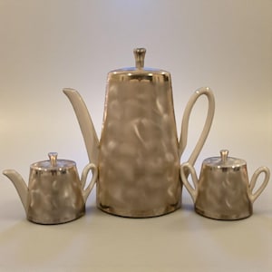 May include: A vintage tea set featuring a large teapot and two smaller serving pieces. The set has a pearlescent finish with silver trim. The teapot has a curved spout and handle, and the serving pieces have lids.