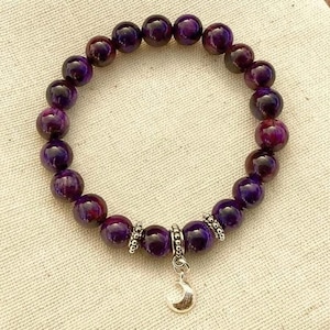 Puede incluir: Una pulsera con piedras preciosas redondas de color púrpura oscuro. La pulsera tiene detalles plateados y un pequeño dije de media luna. Las piedras preciosas tienen un aspecto veteado, con toques de rojo y púrpura más oscuro.