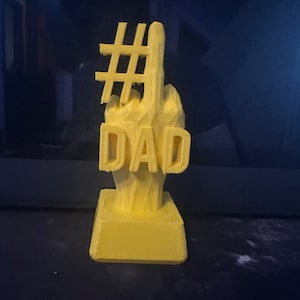 May include: A yellow 3D printed figurine of a hand giving the number one sign with the word "DAD" below it and a hashtag symbol above the hand.