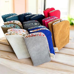 Peut inclure: Une collection de pochettes en velours côtelé de différentes couleurs, dont blanc, gris, bleu et jaune moutarde. Certaines pochettes présentent des doublures à motifs floraux. Les pochettes sont empilées, mettant en valeur leurs différents motifs et textures.