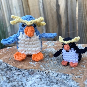 May include: Two crocheted penguin figurines. One is blue, white, and orange, the other is black, white, and orange. Both have yellow accents on their heads and are standing on a rock.