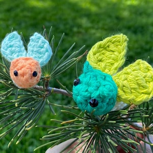 May include: Two handmade crochet insects, one orange with blue wings and the other teal with yellow wings, are perched on a green pine branch. The insects have black bead eyes against a blurred green background.