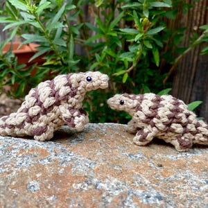 May include: Two handmade crochet pangolins in a natural color palette of beige and brown, with blue bead eyes. The larger pangolin is approximately 6 inches long, and the smaller one is about 4 inches long. They are displayed on a rock.
