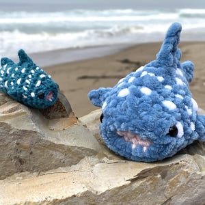 May include: Two crocheted whale shark plushies, one blue with white spots and one teal with white spots, are sitting on a rock near the ocean.