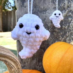 Puede incluir: Dos adornos de fantasmas de crochet blanco con ojos negros y bocas sonrientes que cuelgan de una cuerda. El fantasma más grande está en primer plano y el fantasma más pequeño está en el fondo. Una calabaza está en la esquina inferior derecha de la imagen.