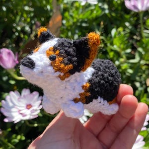 May include: A crocheted stuffed toy dog with black, white, and orange fur. The dog is sitting in a person's hand.
