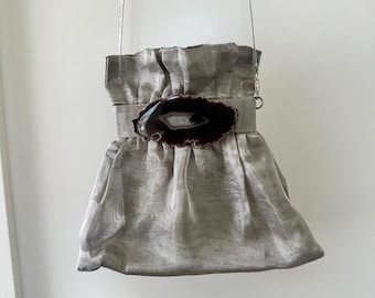 Puede incluir: Un bolso gris plateado con un diseño fruncido y un cierre decorativo de piedra de ágata. El bolso tiene una correa de cadena plateada y una parte superior con volantes. La piedra de ágata es marrón y blanca con un acabado pulido.