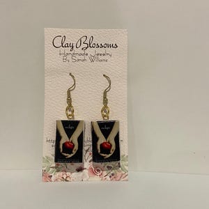 Puede incluir: Un par de pendientes colgantes con réplicas en miniatura de la portada del libro Twilight. Los pendientes están hechos de arcilla polimérica y tienen ganchos de color dorado.