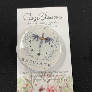 Op de afbeelding: Een ronde, witte pop-up telefoonhouder met een zwart en goudkleurig drakenontwerp, met de woorden "BASGIATH" en "WAR COLLEGE". De houder staat op een witte kaart met de tekst "Clay Blossoms Handmade Jewelry".