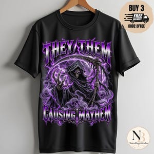 Puede incluir: Una camiseta negra de manga corta con un estampado gráfico de una figura con capucha y guadaña, rodeada de rayos púrpuras y calaveras. El texto "THEY/THEM" aparece en la parte superior y "CAUSING MAYHEM" en la parte inferior en color púrpura.