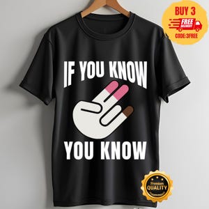 Könnte beinhalten: Ein schwarzes kurzärmeliges T-Shirt mit dem weißen Text IF YOU KNOW YOU KNOW, der mittig über und unter einer Grafik einer Hand mit einer Zwei-Finger-Geste platziert ist. Die Handillustration hat einen weißen Hautton, rosa Fingerspitzen an den beiden erhobenen Fingern und eine braune Fingerspitze am Daumen.