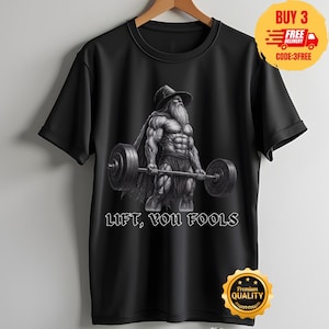 Könnte beinhalten: Ein schwarzes T-Shirt mit kurzen Ärmeln und einer Grafik eines muskulösen, bärtigen Mannes mit breitkrempigem Hut und Umhang, der eine Langhantel hebt. Der Text LIFT, YOU FOOLS ist in weißen Großbuchstaben unter der Grafik aufgedruckt.