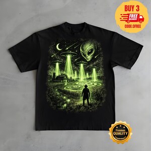 May include: A black t-shirt featuring a graphic print of a glowing green alien scene. The design shows multiple flying saucers hovering over a farm field with a person standing in the foreground, a large alien face in the sky, and a crescent moon.