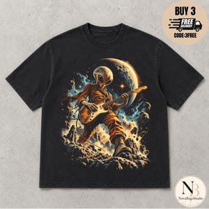Op de afbeelding: Een zwart t-shirt met korte mouwen en een grafische print van een alien in een oranje jumpsuit die op een elektrische gitaar speelt. De achtergrond toont een rotsachtig maanlandschap met een grote maan, kosmische wolken en zwevende asteroïden.
