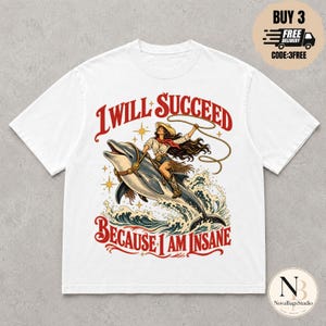 May include: A white short-sleeved t-shirt featuring a graphic of a person in a cowboy hat and boots riding a dolphin while holding a lasso. The text reads I WILL SUCCEED BECAUSE I AM INSANE in red lettering, surrounded by stars and ocean waves.