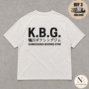 Pode incluir: Uma t-shirt de manga curta em branco sujo com texto preto nas costas. A impressão apresenta as letras K.B.G. numa fonte grande e desgastada, seguidas de caracteres japoneses e o texto KAMOGAWA BOXING GYM numa fonte sem serifa limpa.