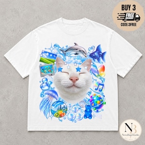 Könnte beinhalten: Ein weißes T-Shirt mit einem Grafikdruck eines lächelnden weißen Katzengesichts, umgeben von blauen Motiven wie einem Delfin, einem Goldfisch, einer Qualle, einem Gummibärchen, einer Discokugel, schwebenden Blasen und kleinen blauen Sternen.